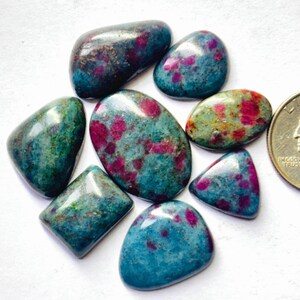 Puede incluir: Una colección de siete piedras preciosas pulidas y lisas, talladas en cabujón, con un color base azul verdoso profundo e inclusiones vibrantes en magenta y rosa, mostradas junto a una moneda para comparar el tamaño.