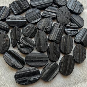 May include: A collection of black tourmaline cabochons, each with a striated pattern. The stones are oval shaped and have a smooth, polished finish.