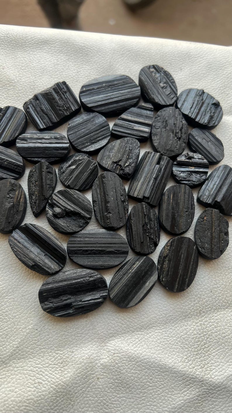 May include: A collection of black tourmaline cabochons, each with a striated pattern. The stones are oval shaped and have a smooth, polished finish.