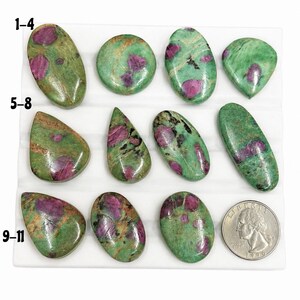 May include: Eleven polished ruby in zoisite cabochons arranged in rows on a white display tray. The stones feature a green base with magenta inclusions and black flecks. A US quarter coin is included for scale next to the bottom row of stones.