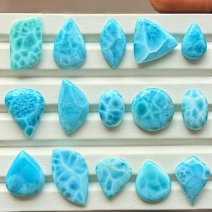 Pode incluir: Uma cole&ccedil;&atilde;o de quinze pedras preciosas Larimar polidas dispostas em tr&ecirc;s filas numa bandeja de exibi&ccedil;&atilde;o branca. Cada pedra apresenta uma cor azul-celeste distinta com padr&otilde;es brancos em forma de teia e v&aacute;rias formas.