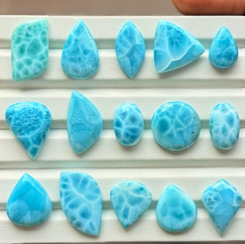 Pode incluir: Uma cole&ccedil;&atilde;o de quinze pedras preciosas Larimar polidas dispostas em tr&ecirc;s filas numa bandeja de exibi&ccedil;&atilde;o branca. Cada pedra apresenta uma cor azul-celeste distinta com padr&otilde;es brancos em forma de teia e v&aacute;rias formas.