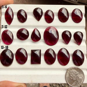 Può includere: Una collezione di diciotto pietre preziose granato rosso scuro lucidate, disposte su un vassoio espositore bianco con tre file etichettate 1-6, 7-12 e 13-18. Le pietre presentano varie forme, tra cui tagli ovali, a goccia e rettangolari.