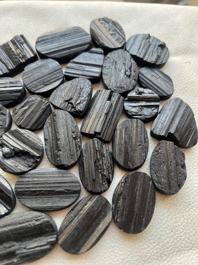 May include: A collection of black tourmaline gemstone cabochons, each with a unique striated pattern. The stones are oval shaped and have a smooth, polished finish.
