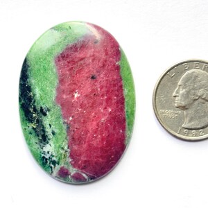 Puede incluir: Una piedra preciosa de zoisita pulida de forma ovalada con una banda central prominente de rubí rosa intenso rodeada de inclusiones minerales de color verde vibrante y negro, junto a una moneda de cuarto de dólar para escala.