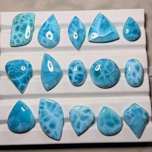 Pode incluir: Uma cole&ccedil;&atilde;o de catorze pedras preciosas Larimar polidas, dispostas em tr&ecirc;s filas numa bandeja de apresenta&ccedil;&atilde;o branca. Cada pedra apresenta uma cor azul-celeste distinta com padr&otilde;es brancos nublados e um acabamento liso e brilhante.
