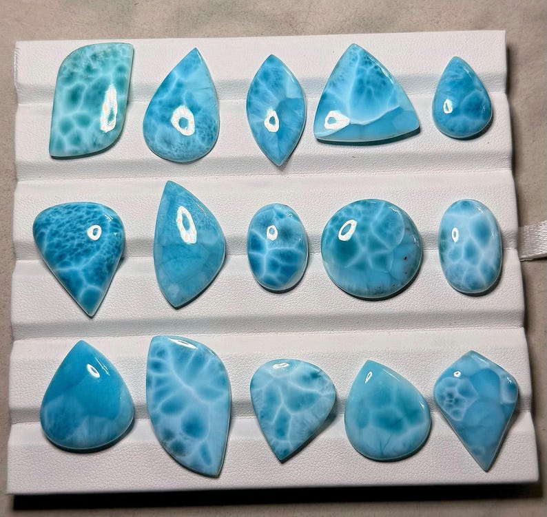 Pode incluir: Uma cole&ccedil;&atilde;o de catorze pedras preciosas Larimar polidas, dispostas em tr&ecirc;s filas numa bandeja de apresenta&ccedil;&atilde;o branca. Cada pedra apresenta uma cor azul-celeste distinta com padr&otilde;es brancos nublados e um acabamento liso e brilhante.
