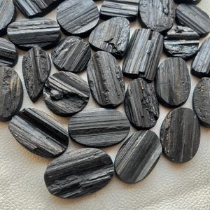 May include: A collection of black tourmaline cabochons, each with a striated pattern. The stones are oval shaped and have a smooth, polished finish.