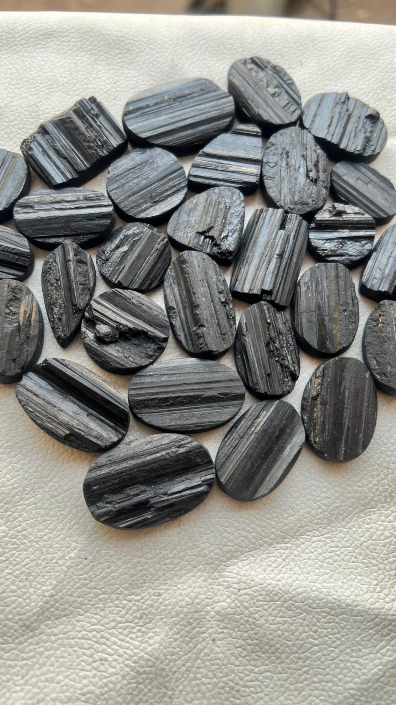 May include: A collection of black tourmaline cabochons, each with a striated pattern. The stones are oval shaped and have a smooth, polished finish.