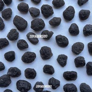 May include: A collection of 30 black obsidian tumbled stones. The stones are smooth and have a shiny, glassy finish. The stones are arranged in a random pattern on a white background. The text "ZOYAGEMS017" appears twice in the image.