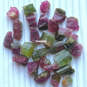 May include: A selection of raw tourmaline gemstones in pink, green, and mixed hues. The rough-cut stones vary in size and shape, showing natural textures and colour variations. The text "zoyagems017" is visible.