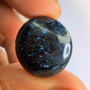 Op de afbeelding: Een ronde, gepolijste cabochon edelsteen met een diepzwarte basiskleur en verspreide, iriserende blauwe insluitingen die het licht vangen. Het oppervlak is glad en reflecterend, met een donker, mineraalrijk uiterlijk met opvallende blauwe vlekjes.