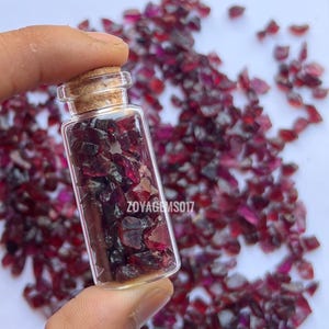 Puede incluir: Una pequeña botella de vidrio transparente llena de piedras preciosas de color rojo oscuro, talladas en bruto. La botella tiene un tapón de corcho y se sostiene sobre un fondo de piedras preciosas dispersas. El texto "ZOYAGEMS017" está impreso.