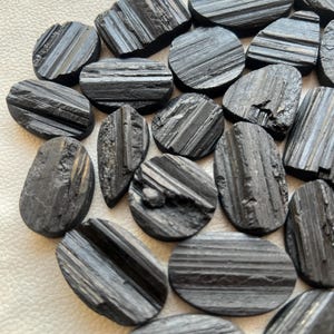 May include: A collection of black, oval-shaped, striated, polished stones. The stones have a rough, natural texture and are arranged in a pile.