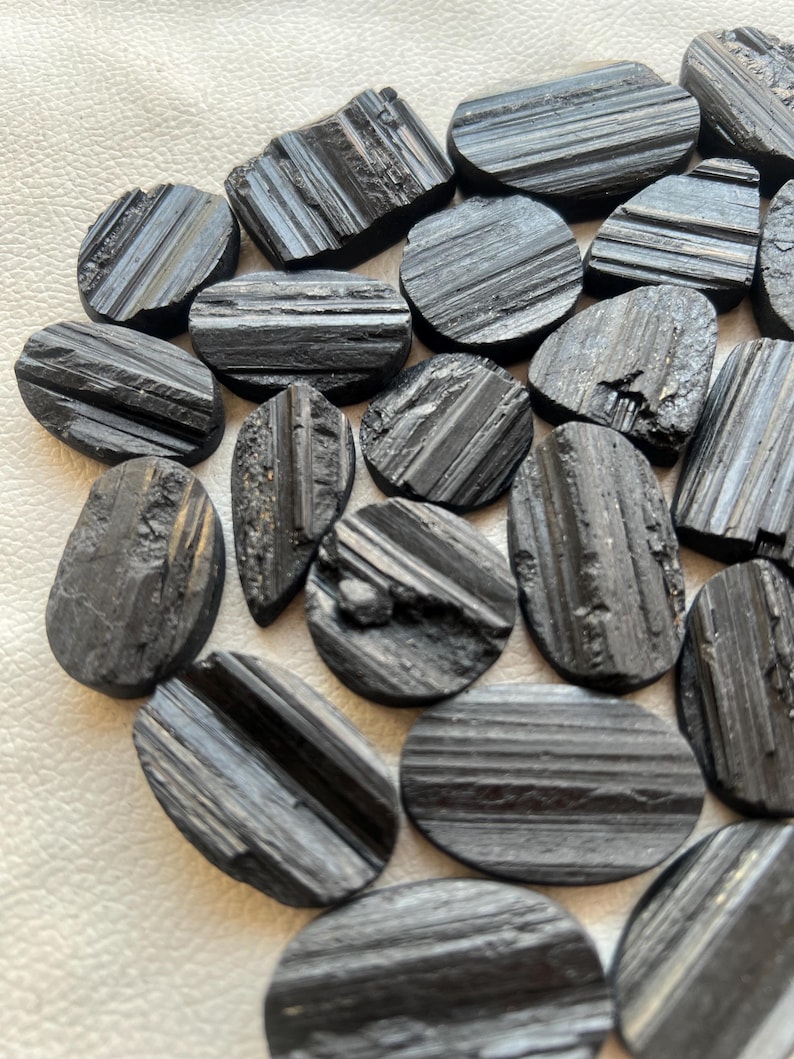 May include: A collection of black, oval-shaped, striated, polished stones. The stones have a rough, natural texture and are arranged in a pile.