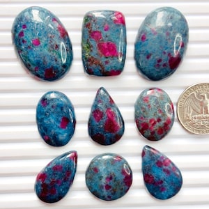 May include: Eight polished cabochon gemstones featuring a blue base with vibrant pink and red inclusions, arranged in three rows. A United States quarter coin is placed on the right side for scale, showing the relative size of the stones.