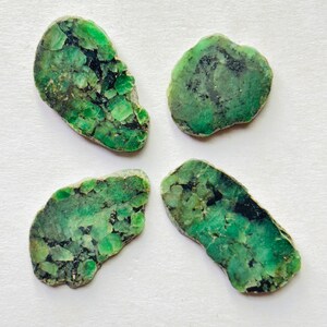 Könnte beinhalten: Vier roh geschnittene Smaragdsteine mit leuchtend grüner Farbe und dunklen Einschlüssen. Die Steine haben unregelmäßige Formen und ein natürliches, unpoliertes Aussehen. Die Smaragde werden auf einer weißen Oberfläche präsentiert.
