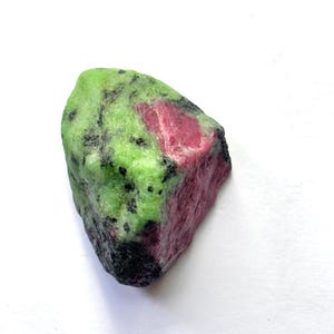 Peut inclure: Un spécimen minéral brut et non poli présentant une matrice de zoïsite vert lime vif avec des inclusions proéminentes de rubis magenta profond et des cristaux de hornblende noirs dispersés, affichant une surface cristalline naturelle.