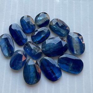 May include: A collection of twelve blue kyanite gemstones, each with a unique, faceted shape. The stones are arranged in a cluster on a white surface.