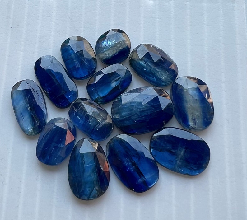 May include: A collection of twelve blue kyanite gemstones, each with a unique, faceted shape. The stones are arranged in a cluster on a white surface.