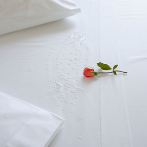 May include: White embroidered bedding with a single orange rose and green stem on top.