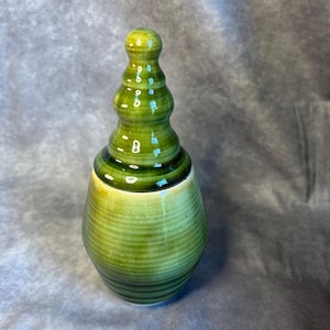 Puede incluir: Un jarrón de cerámica verde con una base bulbosa y un cuello alargado escalonado. El jarrón tiene crestas horizontales y un acabado brillante, con un degradado de color de amarillo claro a verde oscuro.
