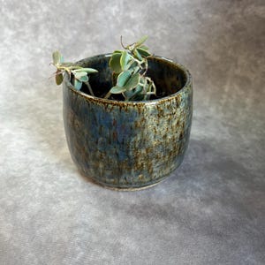 Może przedstawiać: Mała, ręcznie robiona ceramiczna doniczka z teksturowanym, nakrapianym niebiesko-brązowym szkliwem. Cylindryczna doniczka zawiera małą roślinę sukulentową z zielonymi liśćmi. Doniczka nadaje się do małych roślin i dodaje odrobinę naturalnego piękna.