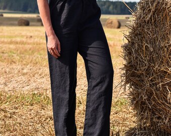 May include: A woman wearing a white off-the-shoulder crop top and black linen pants stands in a field of hay bales.