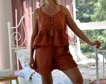 Puede incluir: Una mujer lleva un conjunto de pijama de lino color óxido con una parte superior abotonada y pantalones cortos. La parte superior tiene un dobladillo con volantes y los pantalones cortos tienen un ribete con volantes. La mujer está de pie en un dormitorio con una cama blanca y ropa de cama rosa.