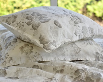 May include: Two white pillowcases with a grey floral pattern. The pillowcases are stacked on top of each other and are lying on a white bedspread with the same floral pattern.