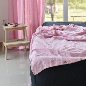 May include: A pink linen bed sheet with a button closure, draped over a black bed with a white wooden step stool in the background.