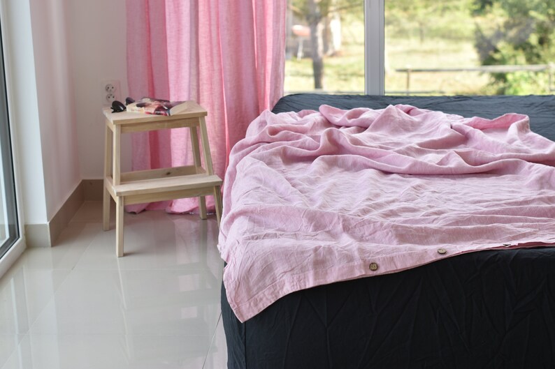 May include: A pink linen bed sheet with a button closure, draped over a black bed with a white wooden step stool in the background.