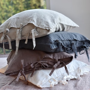 May include: Four linen pillowcases stacked on top of each other. The top pillowcase is light gray, the second is dark gray, the third is brown, and the bottom is white. All pillowcases have ties at the corners.