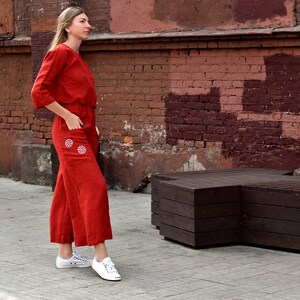 May include: A woman wearing a red linen jumpsuit with white embroidered details. She is standing in front of a red brick wall and a wooden bench.
