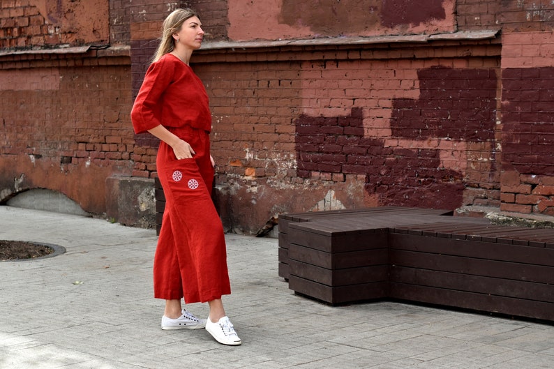May include: A woman wearing a red linen jumpsuit with white embroidered details. She is standing in front of a red brick wall and a wooden bench.