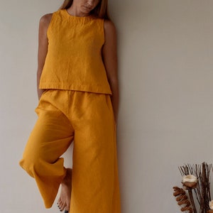 Puede incluir: Conjunto de dos piezas de lino amarillo mostaza. El conjunto incluye un top sin mangas y pantalones de pernera ancha. La tela parece ligera y transpirable. El atuendo es de color liso y tiene un estilo casual y relajado.