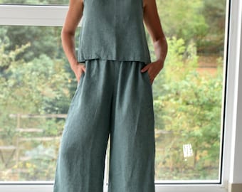 以下が含まれることがあります： ノースリーブトップとワイドレッグパンツの組み合わせた、ティール色のリネンジャンプスーツ。モデルは白いスニーカーを履いています。