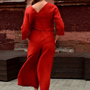 May include: A woman wearing a red linen jumpsuit with a V-neck top and wide-leg pants. She is standing in front of a brick wall and wearing white sneakers.