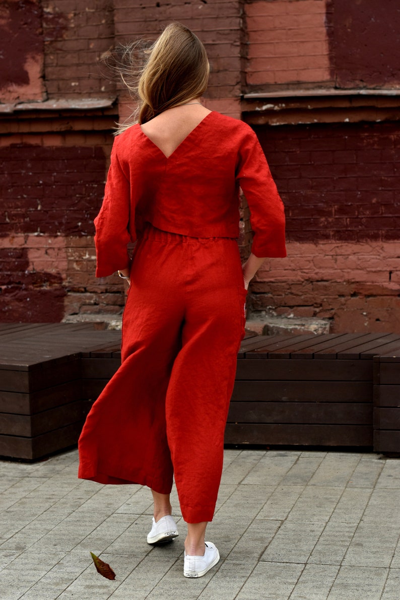 May include: A woman wearing a red linen jumpsuit with a V-neck top and wide-leg pants. She is standing in front of a brick wall and wearing white sneakers.