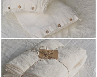 Puede incluir: Un juego de fundas de almohada de lino blanco con botones de madera. Las fundas de almohada están dobladas y atadas con cuerda. El paquete incluye una pequeña etiqueta con el texto "100% Lino" y "Hecho en Europa".