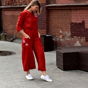 May include: A woman wearing a red linen jumpsuit with embroidered details. The jumpsuit has a cropped top and wide-leg pants. She is wearing white sneakers.