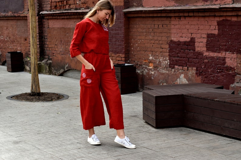 May include: A woman wearing a red linen jumpsuit with embroidered details. The jumpsuit has a cropped top and wide-leg pants. She is wearing white sneakers.