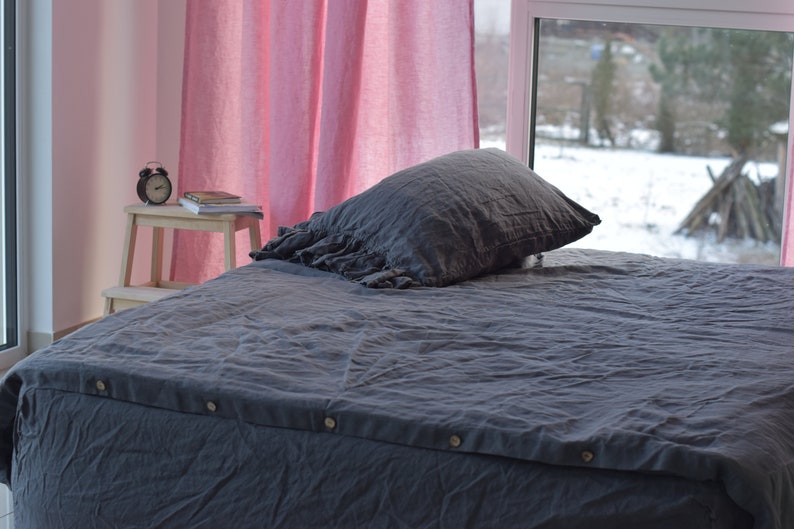 May include: A gray linen duvet cover with a ruffled pillow on a bed. The bed is in a room with pink curtains and a window overlooking a snowy backyard.