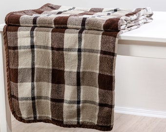 May include: A brown and beige plaid throw blanket with a fringed edge. The blanket is folded and draped over a white table.