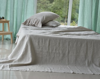 Puede incluir: Una sábana de cama de lino gris claro con un aspecto natural y texturado. La sábana está drapeada sobre una cama con dos almohadas, una de ellas con un borde rizado.