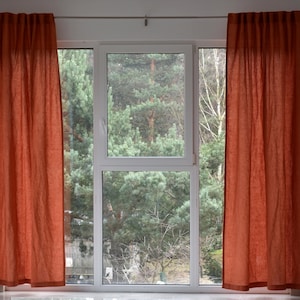 May include: A window with two rust-coloured linen curtains, a white frame, and a view of green trees outside.