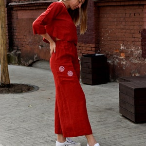 May include: A woman wearing a red linen pantsuit with white embroidered flowers on the pockets. She is wearing white sneakers.