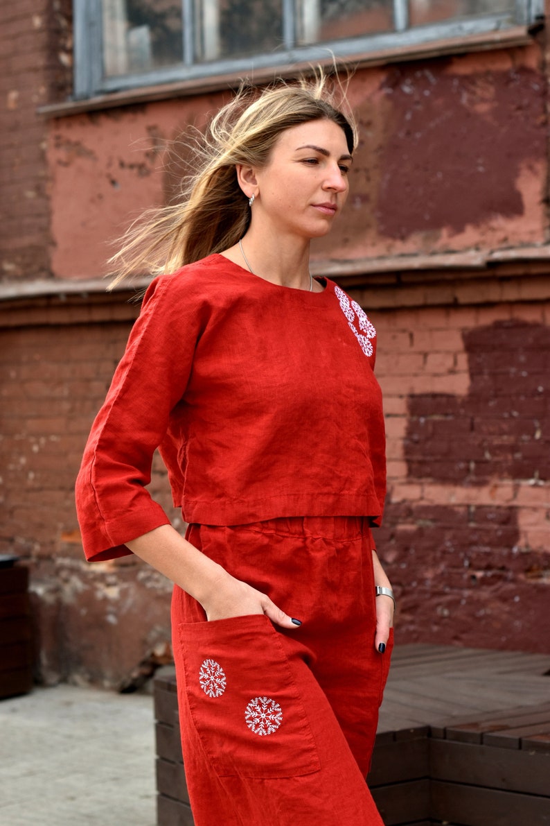 May include: A red linen jumpsuit with white embroidered snowflakes on the top and pockets. The jumpsuit has a cropped top and a wide leg.