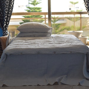 May include: A gray linen bedspread with two white linen pillows on a bed. The bed is in a room with blue walls and a window with curtains.