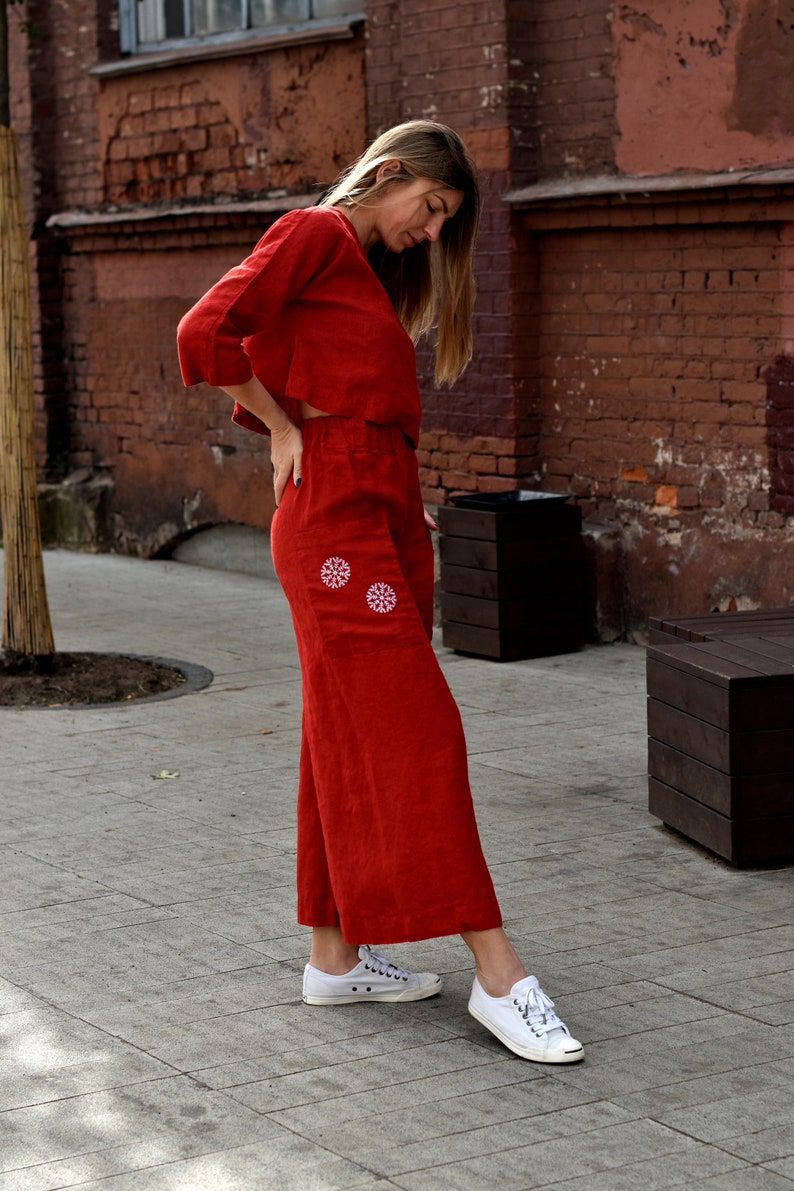 May include: A woman wearing a red linen pantsuit with white embroidered flowers on the pockets. She is wearing white sneakers.
